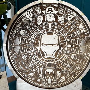 May include: A wooden round plaque with a detailed laser-engraved design featuring various superhero characters from Marvel Comics. The design includes a central image of Iron Man's mask, surrounded by other characters like Captain America, Spider-Man, and Thor.