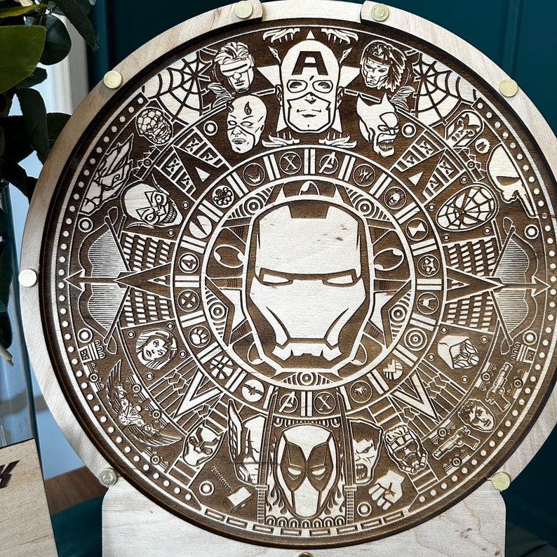 May include: A wooden round plaque with a detailed laser-engraved design featuring various superhero characters from Marvel Comics. The design includes a central image of Iron Man's mask, surrounded by other characters like Captain America, Spider-Man, and Thor.