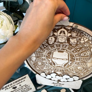 May include: A wooden guest book with a circular design featuring Marvel characters, including Iron Man, Captain America, and Spider-Man. The book is decorated with small wooden hearts and the text "GUESTBOOK PLEASE SIGN A &" and "AUAM & BECWYW 29.08.2021".
