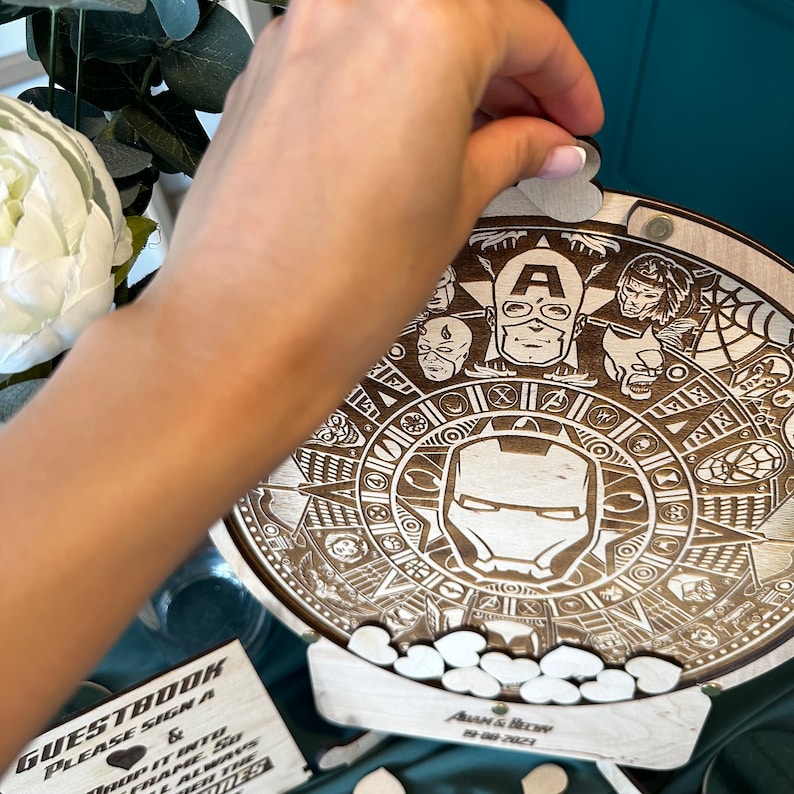May include: A wooden guest book with a circular design featuring Marvel characters, including Iron Man, Captain America, and Spider-Man. The book is decorated with small wooden hearts and the text "GUESTBOOK PLEASE SIGN A &" and "AUAM & BECWYW 29.08.2021".