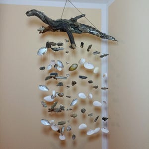 May include: A white and brown seashell wind chime made from driftwood and seashells. The wind chime is hanging from a ceiling and has a rustic look.