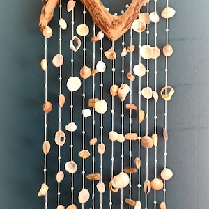May include: A coastal-themed wall hanging featuring a curved piece of driftwood adorned with strings of seashells and small beads. The shells vary in shape and color, creating a natural, textured aesthetic. The piece hangs from a jute rope.
