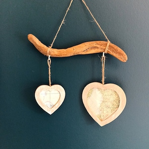 Könnte beinhalten: Rustikale Wanddekoration mit einem natürlichen Ast und zwei herzförmigen Rahmen. Die Rahmen, die an Schnüren hängen, enthalten kartenartige Bilder. Der Hintergrund ist in einem Teal-Ton gehalten.