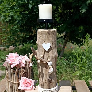 May include: A driftwood candle holder featuring a white heart ornament, wooden beads, and a metal key charm. The pillar sits on a rectangular wooden base wrapped with white rope, topped with a black metal candle cup and a white pillar candle.