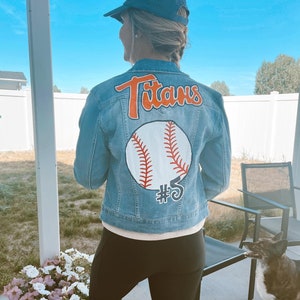 Könnte beinhalten: Eine blaue Jeansjacke mit einem Baseball-Motiv und dem Wort "Titans" auf der Rückseite. Der Baseball ist weiß mit roten Nähten und einem schwarzen Umriss. Die Nummer "#5" ist unter dem Baseball gemalt.