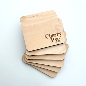 Könnte beinhalten: Ein Stapel quadratischer Holzuntersetzer mit abgerundeten Ecken. Die Untersetzer haben eine helle, natürliche Holzfarbe mit sichtbarer Holzmaserung. Die Worte "Cherry Pye Designs" sind auf dem obersten Untersetzer eingraviert.