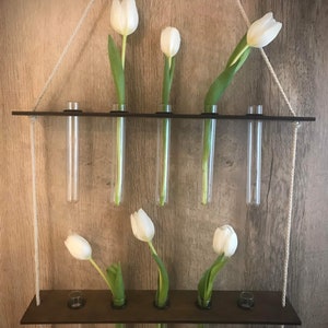 May include: A wooden wall-mounted flower vase with six clear glass tubes. The vase is hanging from a white rope and has white tulips in the tubes.