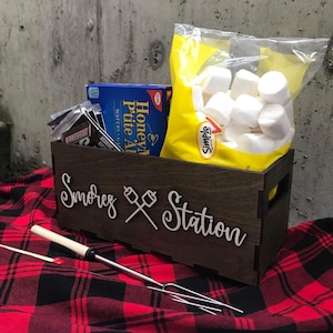 May include: A wooden box with the words "S'mores Station" engraved on the front. The box contains a bag of marshmallows, chocolate bars, and graham crackers. There are also two metal skewers for roasting marshmallows.