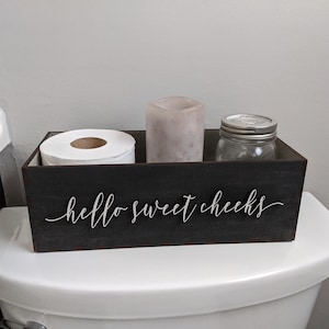 Op de afbeelding: Een zwarte houten doos met de tekst "hello sweet cheeks" aan de voorkant. De doos bevat een rol toiletpapier, een kaars en een glazen pot met deksel.
