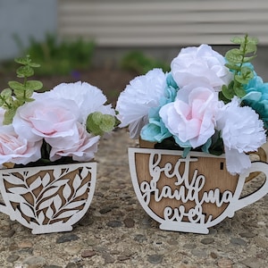 Puede incluir: Dos macetas de madera con forma de taza de té pintadas de blanco con diseños florales. Una maceta dice "mejor abuela de todas" y la otra tiene un patrón de hojas. Ambas macetas están llenas de flores artificiales rosas y azules.