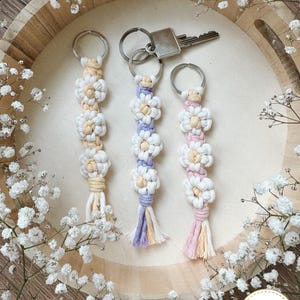 Könnte beinhalten: Drei Makramee-Schlüsselanhänger mit weißen und pastellfarbenen Blumenmotiven. Jeder Anhänger hat einen Metallring, wobei einer einen silbernen Schlüssel hält. Die Blumen bestehen aus cremefarbenen, lavendelfarbenen und rosafarbenen Kordeln.