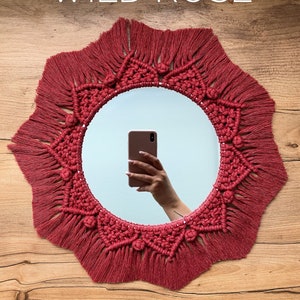 May include: A red macrame mirror with a starburst design and fringe. The mirror is hanging on a wooden wall.
