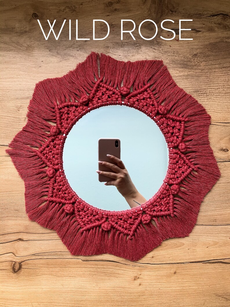 May include: A red macrame mirror with a starburst design and fringe. The mirror is hanging on a wooden wall.