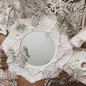 May include: A white macrame mirror with a starburst design and fringe. The mirror is surrounded by white flowers and a wooden background.
