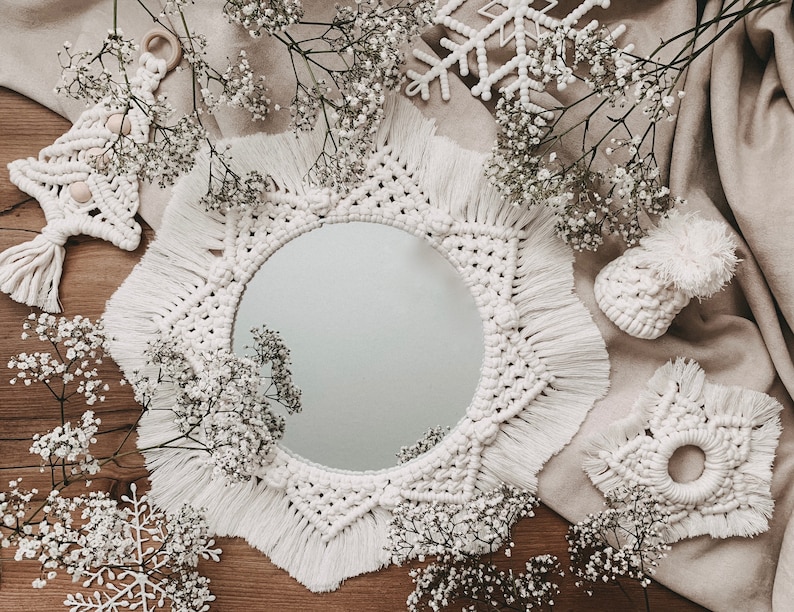 May include: A white macrame mirror with a starburst design and fringe. The mirror is surrounded by white flowers and a wooden background.