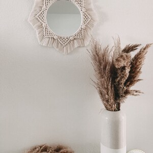 May include: A white macrame mirror with a starburst design hangs on a white wall. A vase with dried pampas grass sits on a table in front of the mirror. The vase is white with a textured finish. Three white candles are in a nest of dried grasses on the table.