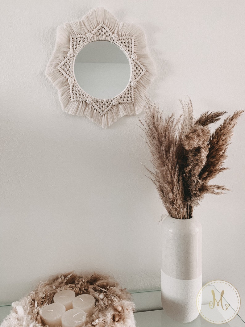 May include: A white macrame mirror with a starburst design hangs on a white wall. A vase with dried pampas grass sits on a table in front of the mirror. The vase is white with a textured finish. Three white candles are in a nest of dried grasses on the table.