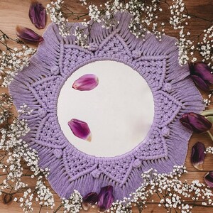 May include: A purple macrame mirror with a round mirror in the center. The mirror is surrounded by white baby's breath flowers and purple flower petals.