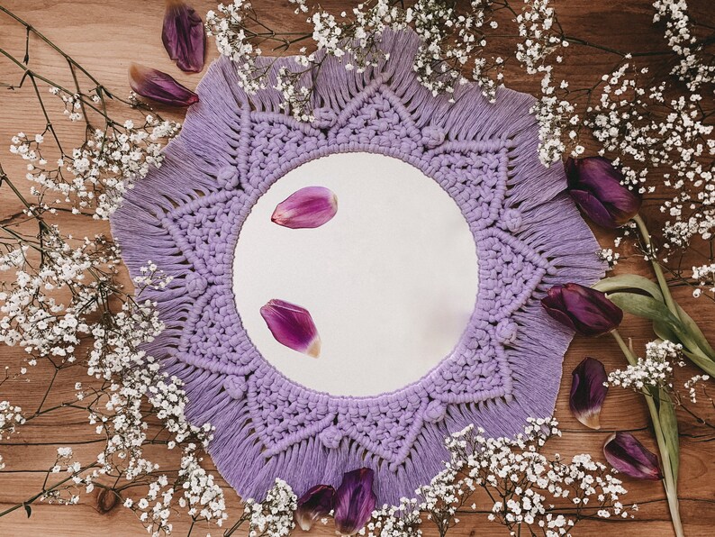 May include: A purple macrame mirror with a round mirror in the center. The mirror is surrounded by white baby's breath flowers and purple flower petals.