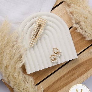 May include: A white ceramic rainbow-shaped display stand with a gold pearl hair clip and a pair of gold pearl earrings. The stand is on a wooden surface with a white fluffy plant.