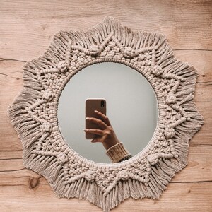 May include: A round mirror with a macrame frame in a natural beige color. The frame has a fringe detail and the mirror is reflecting a hand holding a smartphone. The word "SAND" is written below the mirror.