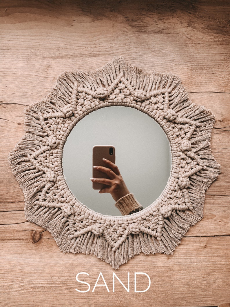 May include: A round mirror with a macrame frame in a natural beige color. The frame has a fringe detail and the mirror is reflecting a hand holding a smartphone. The word "SAND" is written below the mirror.