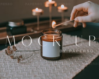 May include: A brown glass candle jar with a white label is being lit with a match. The candle is on a woven placemat with a wooden tray and other candles in the background. The text "MOCK-UP BY PAPERSO" is visible in the image.