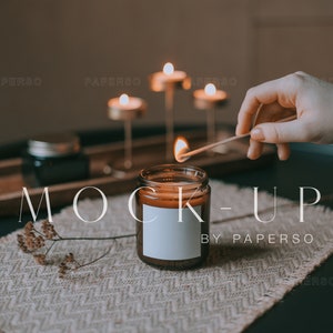 May include: A brown glass candle jar with a white label is being lit with a match. The candle is on a woven placemat with a wooden tray and other candles in the background. The text "MOCK-UP BY PAPERSO" is visible in the image.