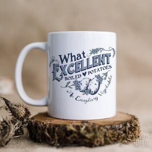 May include: White ceramic mug with the text "What Excellent Boiled Potatoes" in a blue and grey vintage font. The mug features an illustration of potatoes and blue floral accents. The mug is on a wooden coaster.