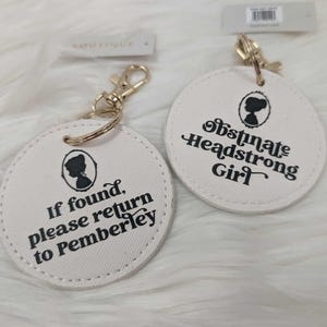 May include: Two white round keyrings with gold-coloured clasps. One keyring reads "If found, please return to Pemberley" and the other says "Obstinate Headstrong Girl" in black text. Each keyring features a silhouette.