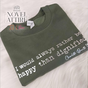May include: An olive green crewneck sweatshirt featuring white text that reads, I would always rather be happy than dignified, followed by the signature Charlotte Bronte. The garment includes a white brand tag at the inner neckline.