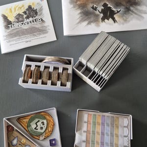 Peut inclure: Des composants de jeu de société, dont un livret de règles intitulé "Nidavellir" et un livret plus petit intitulé "Thingvellir". L'image montre également des porte-cartes, des cartes et des pièces de jeu de différentes couleurs et formes, le tout disposé sur une surface grise.