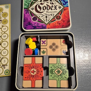 May include: Open board game box with the title "Codex Naturalis" on the lid. Inside, there are game pieces, including wooden tokens in red, yellow, and blue, and illustrated cards in a gray insert.