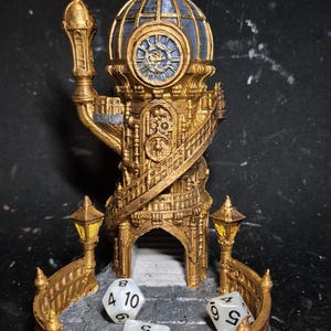 Peut inclure: Une tour d'horloge miniature dorée détaillée avec un dôme bleu et un cadran d'horloge. La tour a un escalier en colimaçon et est entourée d'une balustrade avec deux petites lanternes. Trois dés sont placés devant la tour.
