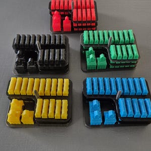 Peut inclure: Vue aérienne de cinq plateaux noirs remplis de pièces de jeu colorées. Les pièces sont rouges, vertes, bleues, jaunes et noires. Les pièces semblent être des bâtiments miniatures et des blocs rectangulaires.
