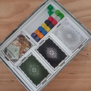 Peut inclure: Boîte de jeu de société ouverte, contenant des cartes aux motifs ornés, des pions colorés et des dés. La boîte est blanche, posée sur un fond en bois.