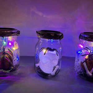May include: Three glass jars with lids decorated with Halloween themes. The jars are filled with colorful fairy lights and contain a ghost, a skull, and a spider.