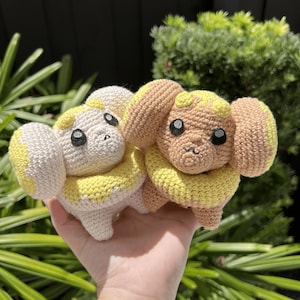 May include: Two small crocheted plush figures featuring large rounded ears, a thick yellow neck collar, and black embroidered eyes. One figure is cream-coloured, while the other is light brown with yellow accents on the ears and forehead.