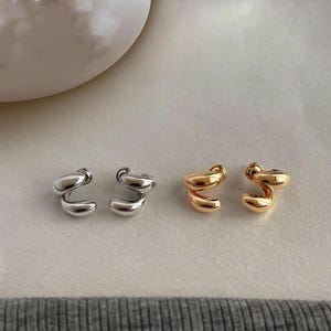 May include: Two pairs of small stud earrings with a smooth, polished finish. One pair is silver-toned and the other pair is gold-toned. Each earring features a curved, twisted shape that loops back on itself, creating a rounded, organic form.