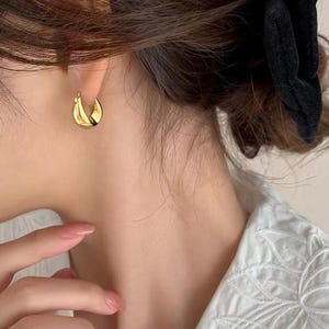 May include: Gold-coloured hoop earrings with a twisted design. The earrings are worn on the ear of a person. The earrings have a shiny finish and reflect light. The person is wearing a white shirt.