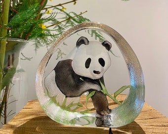 May include: A clear glass sculpture of a panda bear holding a bamboo stalk. The panda is surrounded by green bamboo leaves. The sculpture is on a wooden surface. The sculpture is signed "DR STEINER"