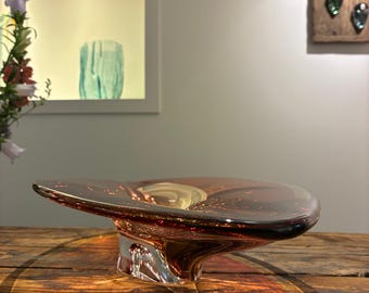 May include: A decorative glass bowl in a reddish-brown colour. The bowl has a wide, shallow design with a flared rim and a clear, sculpted base. The bowl is displayed on a wooden surface, highlighting its unique shape and colour.