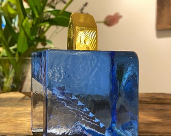 May include: A blue glass bottle stopper with a gold top. The bottle stopper is square shaped and has a textured surface.