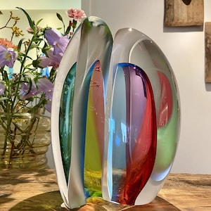 Może przedstawiać: Kolorowa rzeźba ze szkła artystycznego o przejrzystym, zakrzywionym kształcie. Rzeźba charakteryzuje się żywymi odcieniami czerwieni, niebieskiego, zieleni i żółci. Element jest prezentowany na drewnianej powierzchni, prezentując swoją artystyczną formę i wariacje kolorystyczne.