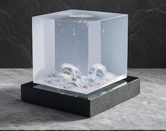 May include: A clear, cube-shaped sculpture with two sculpted faces partially submerged in a gray, textured material. The cube sits on a dark gray rectangular base, creating a modern art piece.