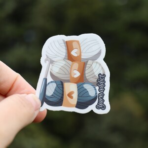 Pu&ograve; includere: Un adesivo con una pila di gomitoli di lana e un uncinetto. I gomitoli di lana sono bianchi, beige e blu. L'adesivo ha il testo "Yarn Addict" sul lato.