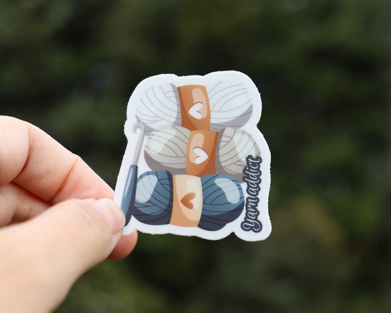 Pu&ograve; includere: Un adesivo con una pila di gomitoli di lana e un uncinetto. I gomitoli di lana sono bianchi, beige e blu. L'adesivo ha il testo "Yarn Addict" sul lato.