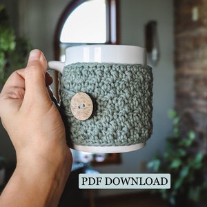 Puede incluir: Un calientavasos de ganchillo de color verde salvia con un botón de madera. El calientavasos está en una taza blanca.