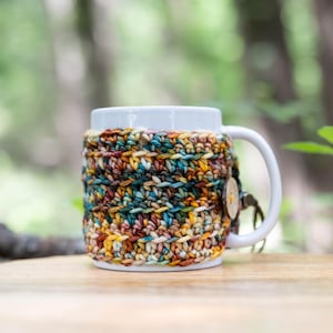 May include: A white ceramic mug featuring a removable, hand-crocheted cosy made of variegated yarn in shades of brown, teal, yellow, and cream. The cosy is secured around the mug with a single round wooden button attached to a small loop.