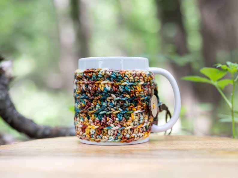 May include: A white ceramic mug featuring a removable, hand-crocheted cosy made of variegated yarn in shades of brown, teal, yellow, and cream. The cosy is secured around the mug with a single round wooden button attached to a small loop.